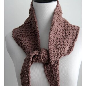 May include: A brown crocheted scarf displayed on a mannequin. The scarf is tied in a knot at the front, with the ends hanging down. The crochet stitch creates a textured appearance. The scarf is a solid color and appears to be handmade.