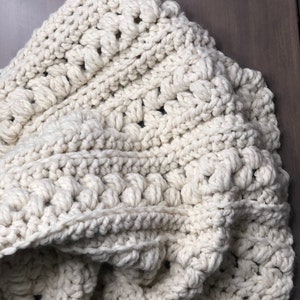 May include: A cream-coloured crocheted blanket with a textured, puffy stitch pattern. The blanket is folded and draped over a wooden surface.