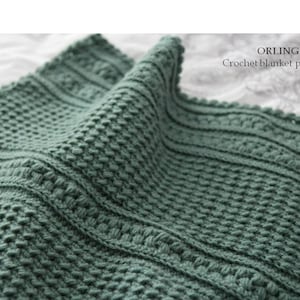 Puede incluir: Primer plano de una manta de ganchillo verde salvia doblada. La manta presenta un patrón de puntadas texturizadas con bandas horizontales y el texto "ORLINGBURY Crochet blanket pattern". La manta está hecha de hilo suave.