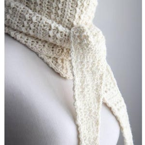 May include: Cream-colored, hand-crocheted scarf with a textured stitch pattern. The scarf is tied in a knot, with two long, pointed ends hanging down. The scarf is displayed on a white background.