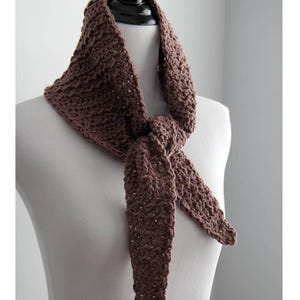 May include: A brown crocheted scarf is displayed on a mannequin. The scarf is knotted at the front, with long, pointed ends. The scarf has a textured pattern and is a solid, warm brown colour. The scarf is a triangular shape.