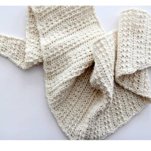 May include: Cream-colored, hand-crocheted scarf. The scarf is made of a soft, textured yarn and features a triangular shape. The crochet stitch creates a dense, warm fabric. The scarf is draped on a white surface, showcasing its intricate design.