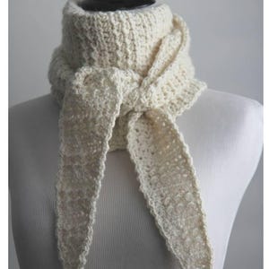 May include: Cream-colored crochet scarf displayed on a mannequin. The scarf is made of thick yarn, with a textured stitch pattern. The scarf is tied in a knot at the front, with long, pointed ends. The text "ORLINGBURY Crochet pattern" is at the bottom.
