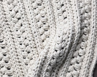 May include: A close-up of a crocheted blanket in a cream color. The blanket is made with a textured stitch that creates a raised pattern.