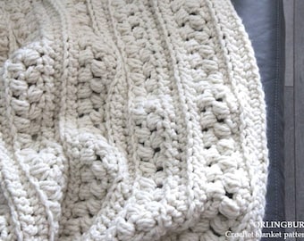 May include: A close-up of a white crocheted blanket with a textured pattern. The blanket is draped over a gray couch.