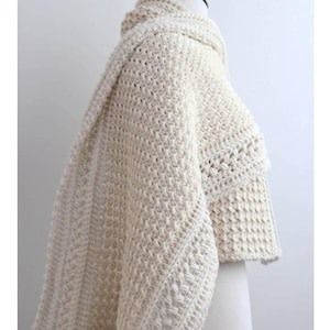 Może przedstawiać: Kremowy, szydełkowy szal ułożony na manekinie. Szal ma wzorzysty ścieg i szeroki, płynny krój. Tekst "ORLINGBURY Crochet shawl pattern" jest widoczny w lewym górnym rogu.