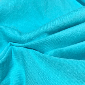 May include: A close-up of a soft, turquoise blue fabric with a smooth texture. The fabric is folded and draped, creating a gentle wave-like pattern.