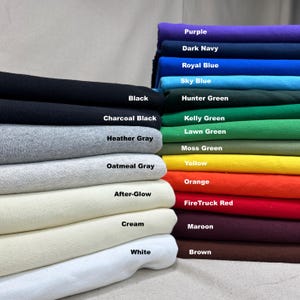 May include: A stack of fabric swatches in various colors, including black, charcoal black, heather gray, and white. Other colors include purple, dark navy, royal blue, sky blue, hunter green, and more. Each swatch is labeled with its color name.