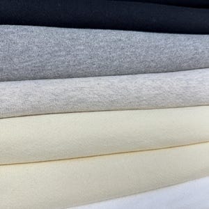 May include: A stack of fabric samples in various colors, including black, gray, cream, and white. The fabric appears to be a soft knit material, suitable for apparel or home decor projects. The samples are neatly folded, showcasing the different shades and textures.