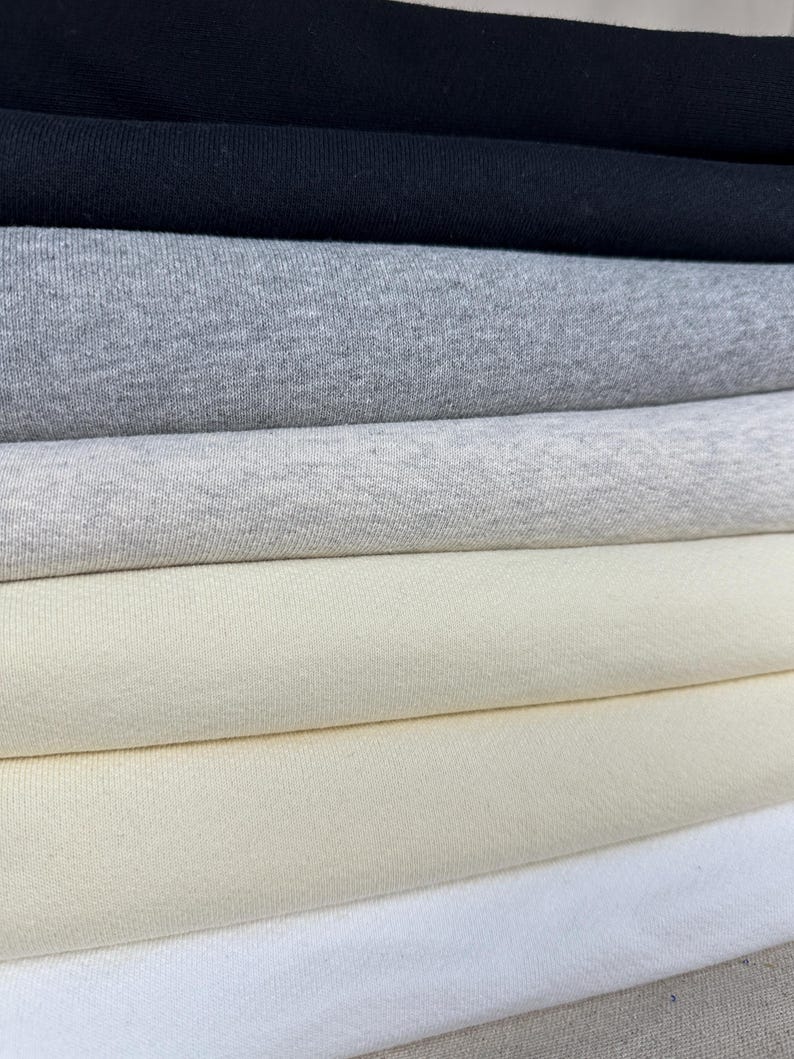 May include: A stack of fabric samples in various colors, including black, gray, cream, and white. The fabric appears to be a soft knit material, suitable for apparel or home decor projects. The samples are neatly folded, showcasing the different shades and textures.