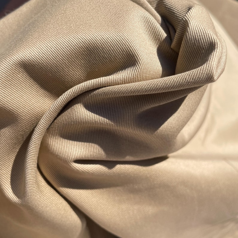 May include: A close-up of a beige fabric with a subtle ribbed texture. The fabric is folded and draped, showcasing its soft and smooth surface.