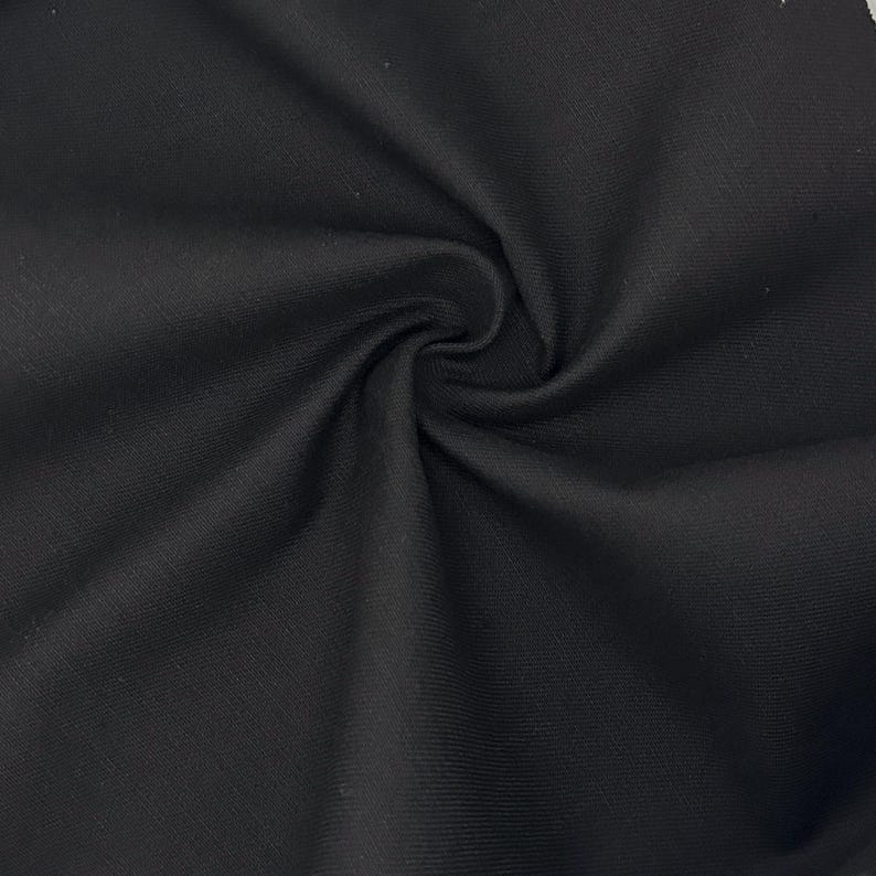 Puede incluir: Una tela negra s&oacute;lida con una textura de tejido fino y uniforme. El material est&aacute; recogido en el centro, creando pliegues suaves y radiantes junto con sombras que resaltan el acabado mate y el color oscuro constante de la superficie.