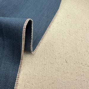 May include: Close-up of denim fabric with a folded edge. The denim is a deep blue colour, and the edge is trimmed with a white and red stitch. The fabric is lying on a beige canvas.