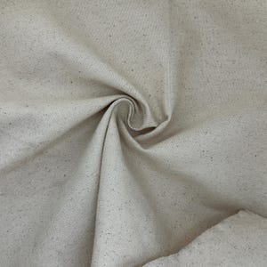 Puede incluir: Un trozo de tela de lona sin blanquear de color natural con una textura tejida visible. El material muestra un tono beige claro con peque&ntilde;as motas m&aacute;s oscuras de materia vegetal org&aacute;nica incrustadas en el tejido, creando un aspecto moteado.