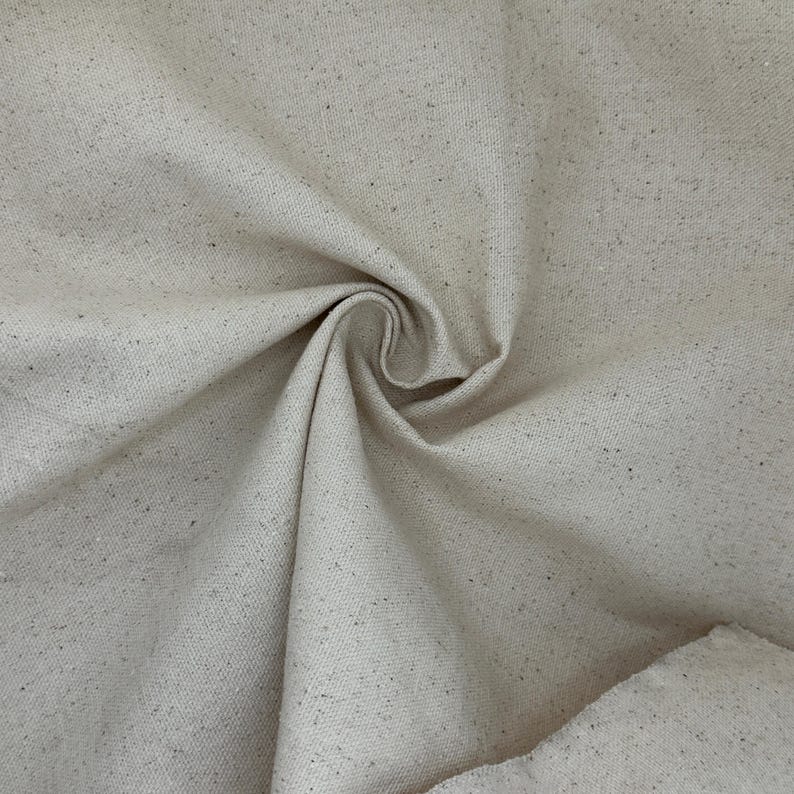 Puede incluir: Un trozo de tela de lona sin blanquear de color natural con una textura tejida visible. El material muestra un tono beige claro con peque&ntilde;as motas m&aacute;s oscuras de materia vegetal org&aacute;nica incrustadas en el tejido, creando un aspecto moteado.