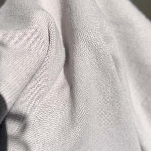 May include: Close-up of a light gray fabric with a textured weave. The fabric is folded and draped, showing the texture and drape of the material.