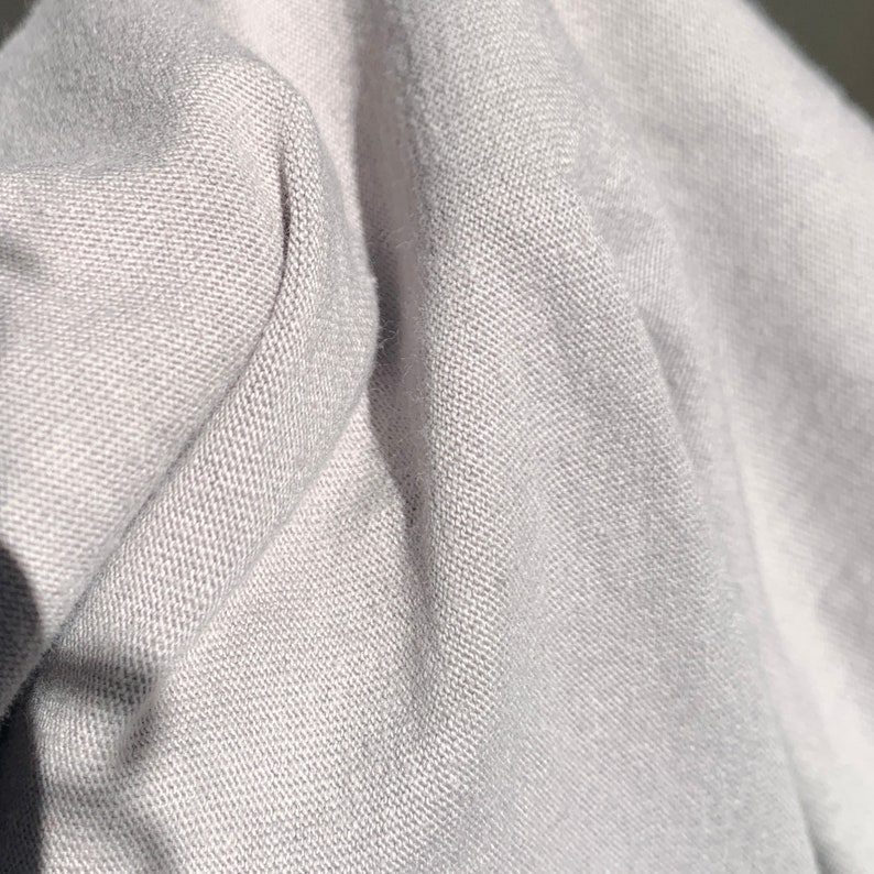 May include: Close-up of a light gray fabric with a textured weave. The fabric is folded and draped, showing the texture and drape of the material.