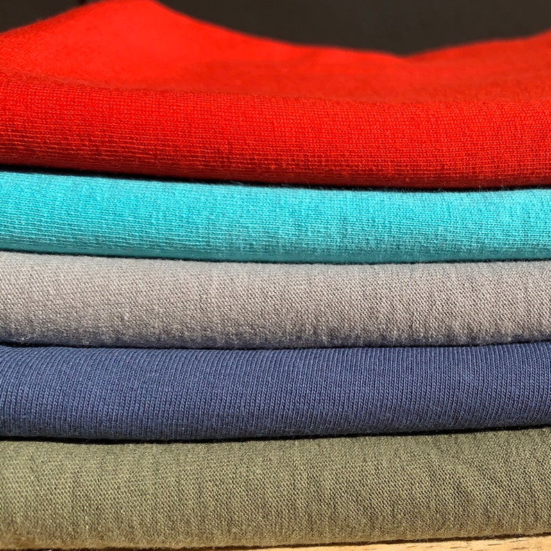 May include: A stack of five folded t-shirts in different colors: red, turquoise, gray, navy blue, and olive green.