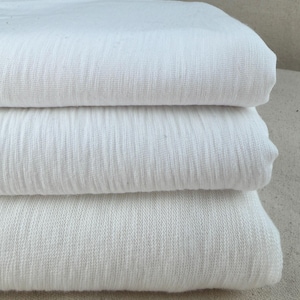 Puede incluir: Una pila de tres piezas de tela de algod&oacute;n blanca dobladas. La tela tiene un aspecto texturizado y ligeramente acanalado. Los pliegues son limpios y la tela parece suave y ligera, adecuada para varios proyectos textiles.