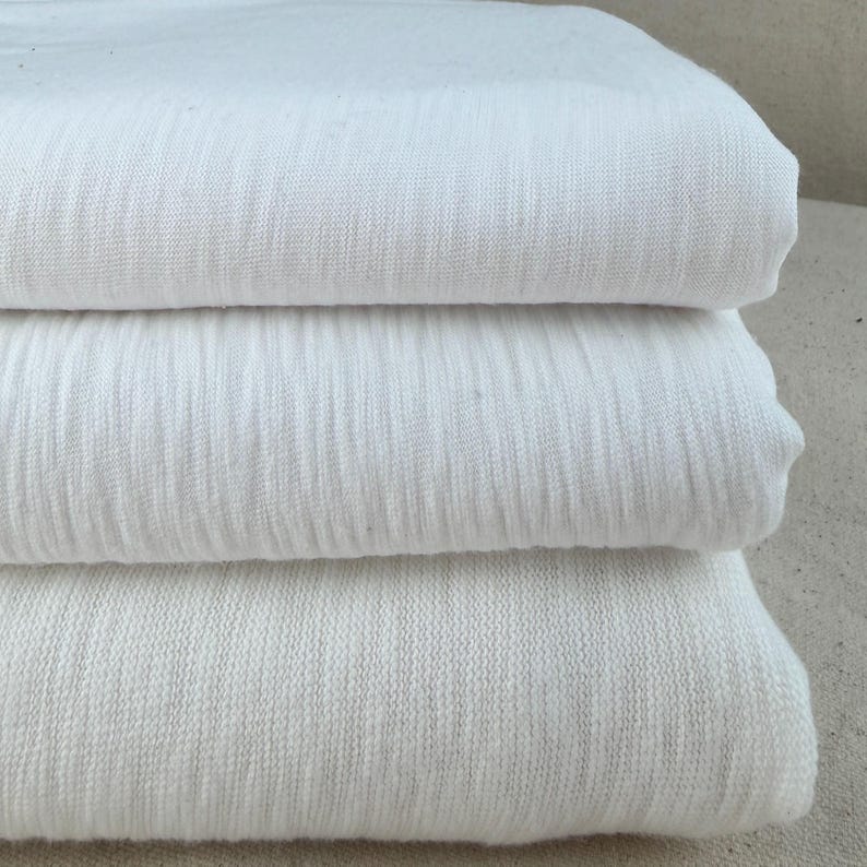 Puede incluir: Una pila de tres piezas de tela de algod&oacute;n blanca dobladas. La tela tiene un aspecto texturizado y ligeramente acanalado. Los pliegues son limpios y la tela parece suave y ligera, adecuada para varios proyectos textiles.