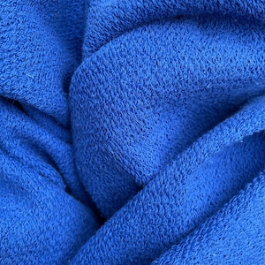 May include: Close-up of a vibrant blue fabric with a textured, woven appearance. The material is folded and draped, creating shadows and highlights that emphasize the texture. The color is a rich, saturated blue.