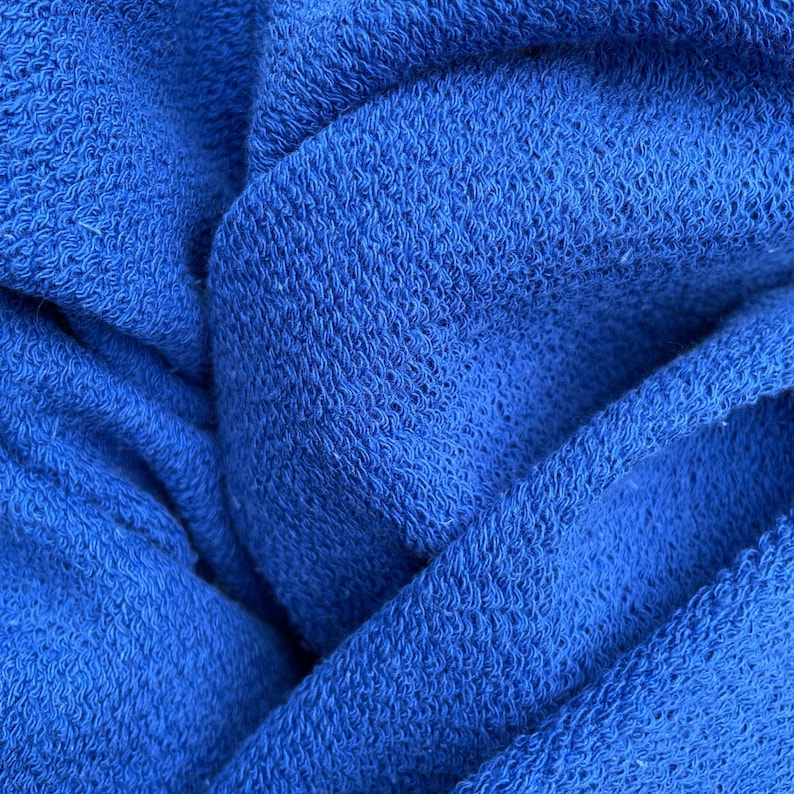 May include: Close-up of a vibrant blue fabric with a textured, woven appearance. The material is folded and draped, creating shadows and highlights that emphasize the texture. The color is a rich, saturated blue.