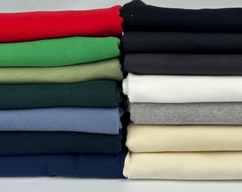 May include: A stack of folded t-shirts in various solid colors including red, green, olive, dark green, light blue, navy, black, white, heather gray, and cream. The garments are neatly layered in two vertical columns against a plain background.