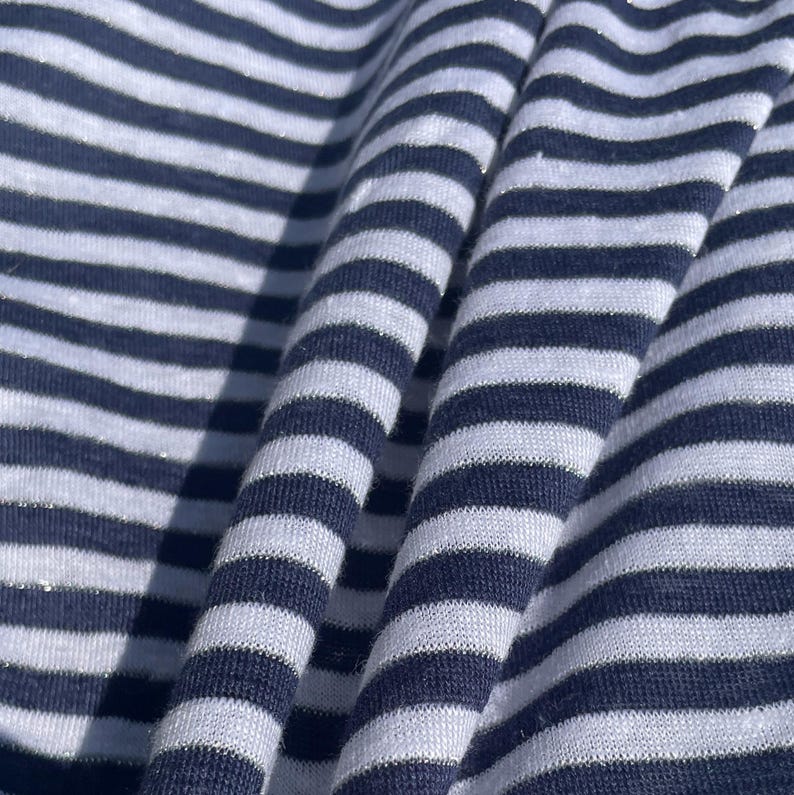 May include: White and navy blue striped fabric with a subtle shimmer. The fabric is folded and draped, showing the texture and pattern.