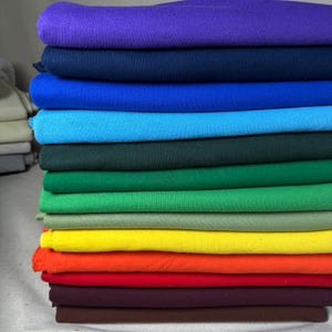 May include: A stack of folded fabric in a variety of colors, including purple, navy, blue, teal, green, yellow, orange, and red. The fabric appears to be a soft material, possibly cotton or fleece. The colors are vibrant and well-saturated.