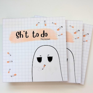 May include: A square notepad with a white grid background featuring a cartoon ghost smoking a cigarette. The text "Sh*t to do" is printed in black over a peach-colored brush stroke, with small cigarette illustrations scattered across the page.