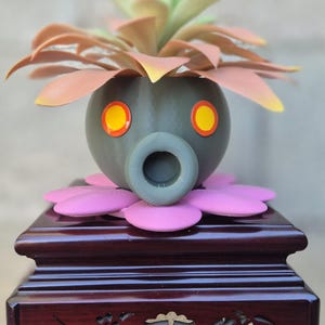 Peut inclure: Un pot de fleurs sphérique gris foncé avec une ouverture circulaire creuse et deux yeux jaune vif bordés d'orange. Le pot repose sur une base en forme de fleur rose et contient une plante succulente artificielle aux feuilles vert clair et pêche.