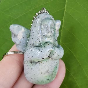 Puede incluir: Una figura de ángel verde claro con detalles moteados, alas y una cadena plateada. El ángel está juntando las manos. La figura está unida a un llavero plateado.