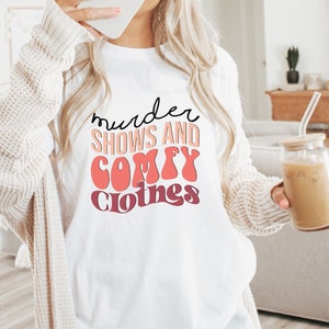 May include: White t-shirt with the text "murder shows and comfy clothes" in a retro font with pink and orange colors.