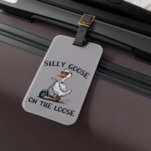 May include: A gray luggage tag with a white goose wearing sunglasses and riding a scooter. The text on the tag reads "Silly Goose on the Loose".