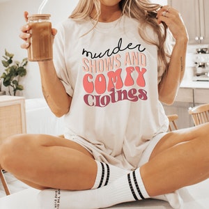 May include: A white t-shirt with the text "murder shows and comfy clothes" in pink and purple lettering.