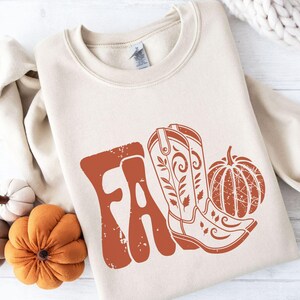 May include: Cream-colored sweatshirt with a rust-colored graphic that says "FA" with a cowboy boot and pumpkin. The boot has floral details. The pumpkin has a textured design. The sweatshirt is folded.