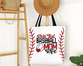 May include: White tote bag with black handles featuring the text "Livin' That Baseball Mom Life" in red and black. The design includes a baseball, a heart, and red stitching accents. A tan hat hangs above the bag.