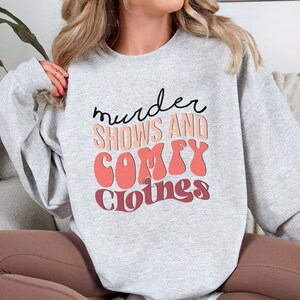 May include: Gray sweatshirt with the text "murder shows and comfy clothes" in a retro font with pink and red accents.