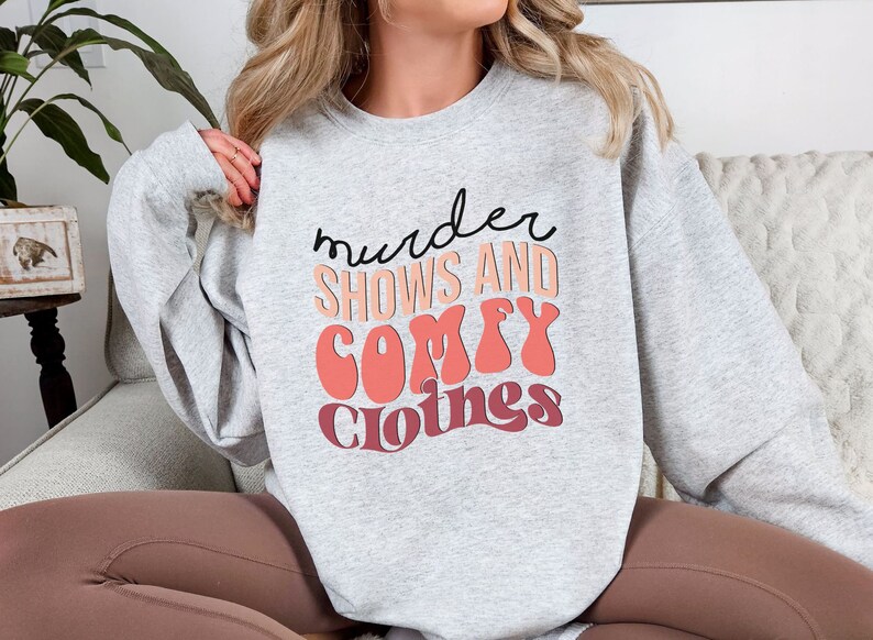 May include: Gray sweatshirt with the text "murder shows and comfy clothes" in a retro font with pink and red accents.