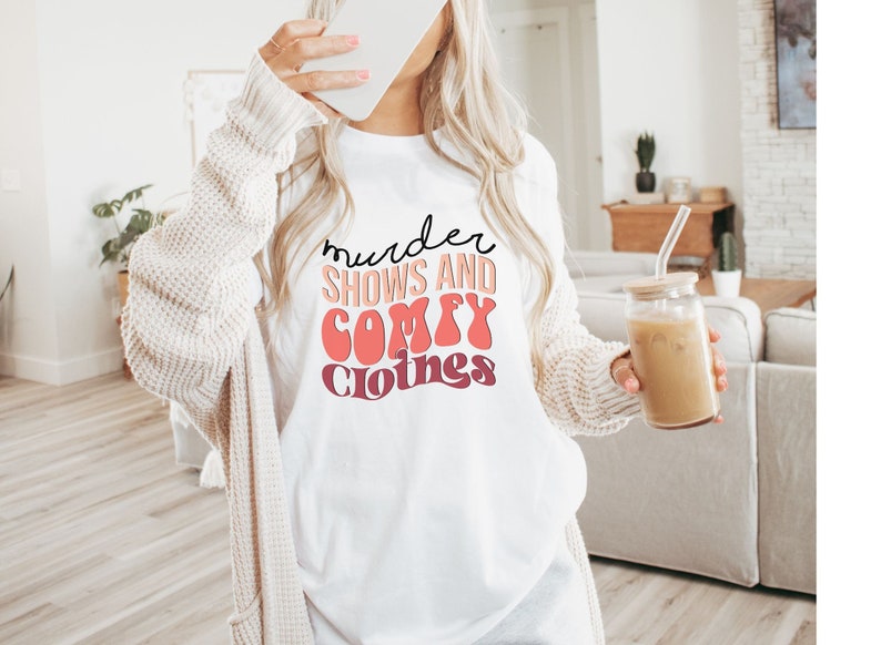 May include: White t-shirt with the text "murder shows and comfy clothes" in a retro font with pink and orange colors.