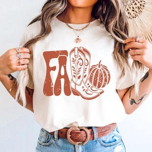 May include: Cream-colored t-shirt with a distressed rust-colored graphic that reads "FA" with a cowboy boot and pumpkin. The shirt is paired with blue jeans and a brown belt with a decorative buckle.