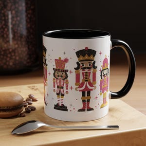 May include: White ceramic mug with a black interior and handle, featuring a festive design of four nutcracker figures in red, pink, and gold. The mug is set on a wooden surface with a spoon and a pastry.