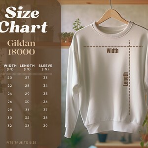 May include: Size chart for Gildan 18000 sweatshirts, showing width, length, and sleeve measurements in inches for sizes S through 4XL.  The chart is displayed on a brown background with a white sweatshirt hanging on a wooden hanger.