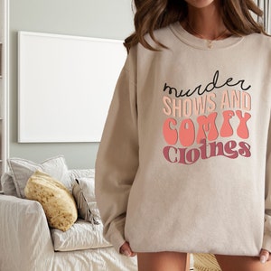May include: A beige sweatshirt with the text "murder shows and comfy clothes" printed in black and pink.
