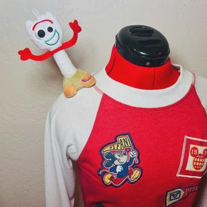 May include: A red and white raglan-style long-sleeved top with Mickey Mouse patches and a Forky plush toy. The top has a red body with white sleeves and a crew neck. Patches include Mickey Mouse with a flag and a globe.