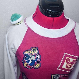 May include: A magenta and white raglan sleeve sweatshirt with Mickey Mouse patches. The patches include Mickey Mouse holding a flag that says "#1 FAN" and a patch with the number "1971". A small green turtle plush is on the shoulder.