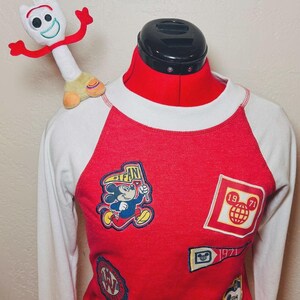 May include: Red and white raglan-sleeve sweatshirt with various Disney patches, including Mickey Mouse and a globe. A plush Forky toy sits on the shoulder. The shirt has a crew neck and long sleeves, ideal for Disney enthusiasts.