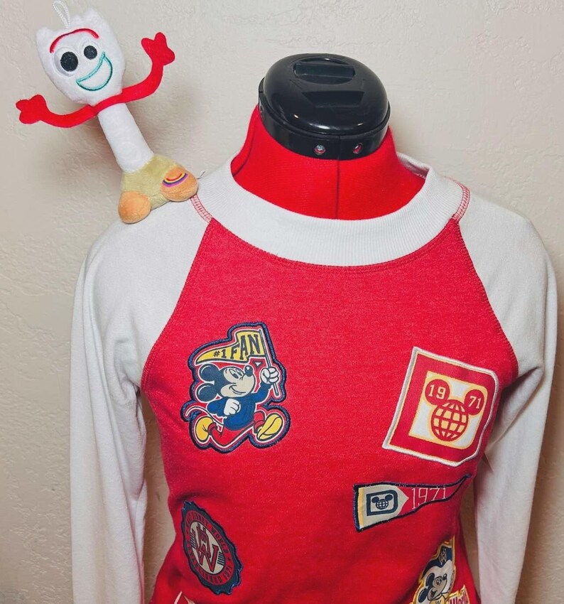 May include: Red and white raglan-sleeve sweatshirt with various Disney patches, including Mickey Mouse and a globe. A plush Forky toy sits on the shoulder. The shirt has a crew neck and long sleeves, ideal for Disney enthusiasts.