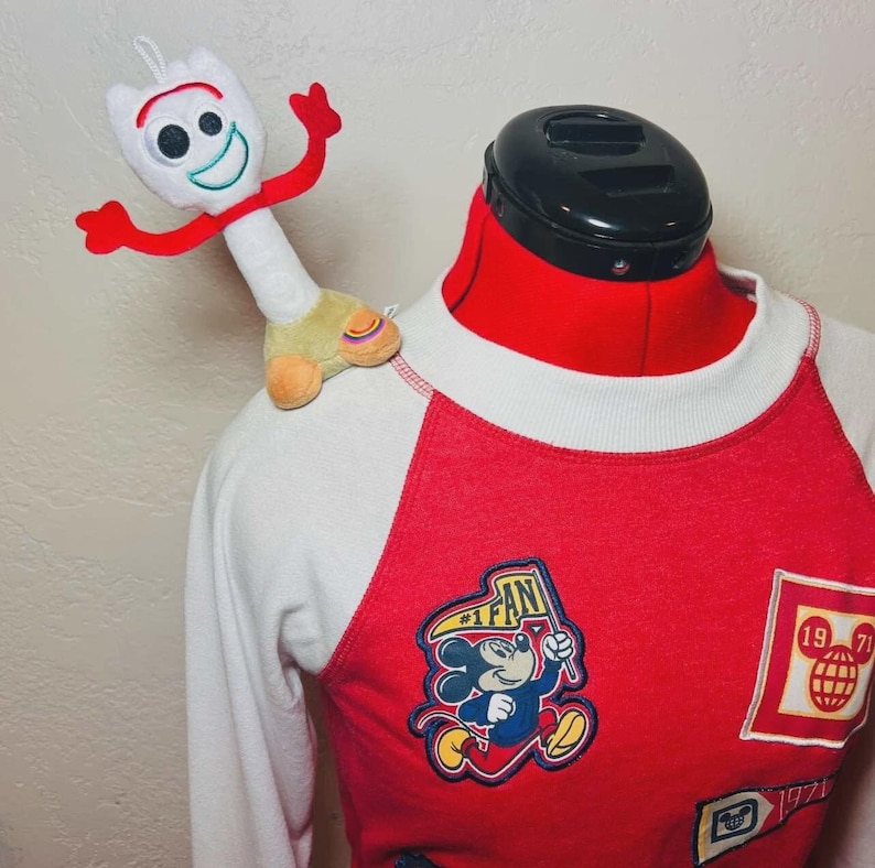 May include: A red and white raglan-style long-sleeved top with Mickey Mouse patches and a Forky plush toy. The top has a red body with white sleeves and a crew neck. Patches include Mickey Mouse with a flag and a globe.