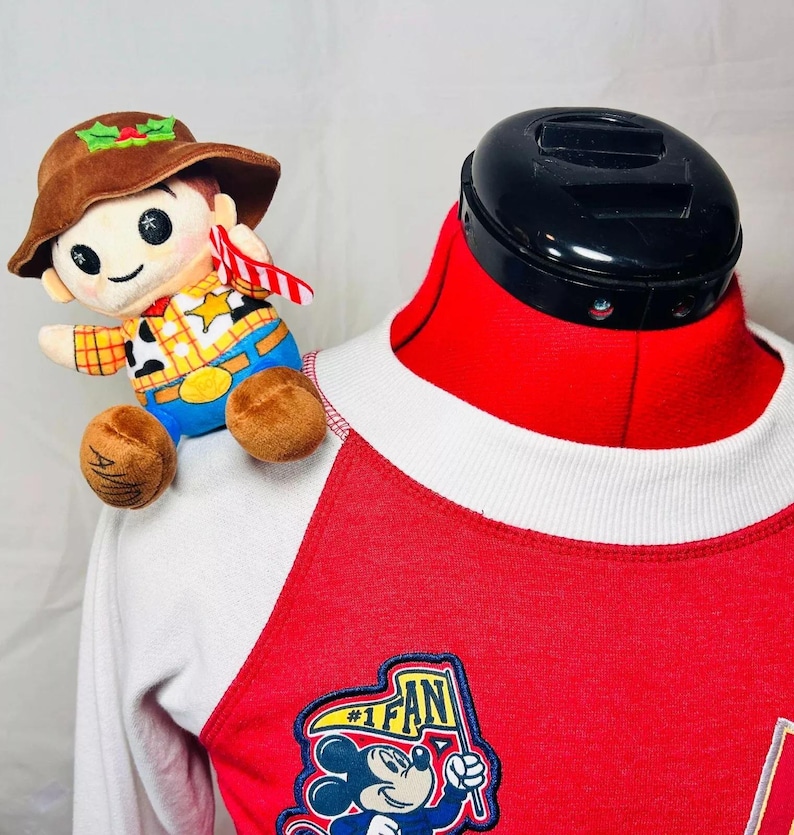 May include: A red and white baseball jersey with a Mickey Mouse #1 Fan patch on the front. A plush toy of Woody from Toy Story is perched on the shoulder of the jersey.